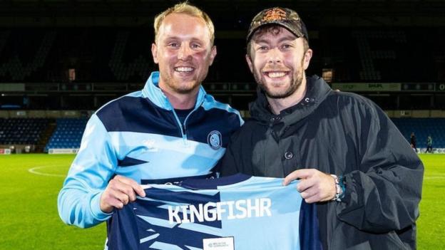 Two men holding a football top that says Kingfishr 1